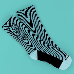 A pair of crew socks with a black and light blue wavy zebra-like pattern, featuring a detailed black graphic on one sock and a white toe and heel, displayed on a solid turquoise background.